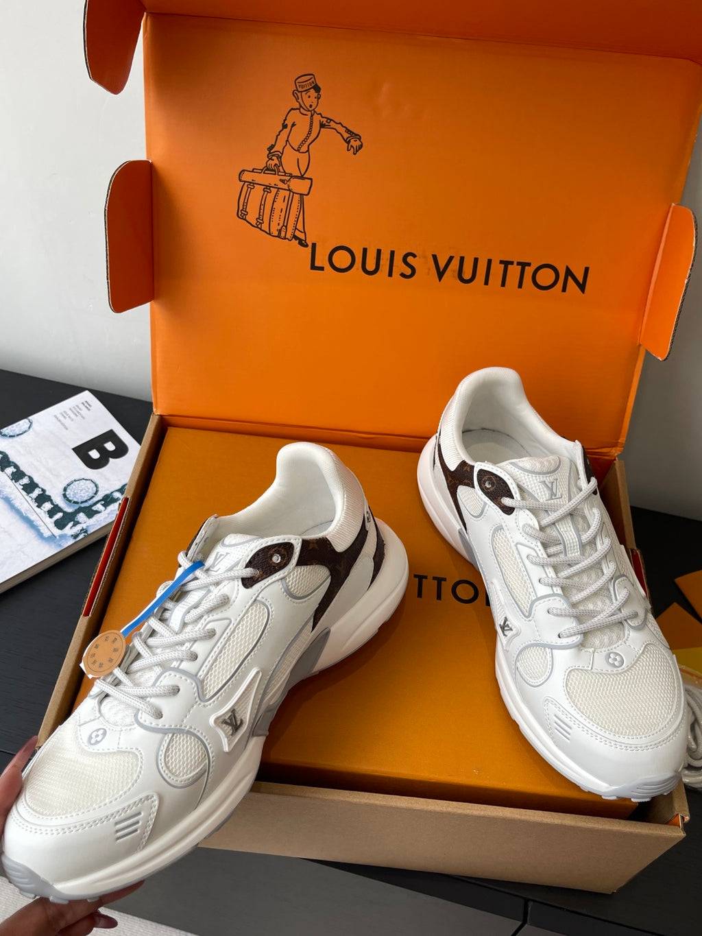 Pair of white lace-up sneakers in an open orange box, angled view with laces visible.