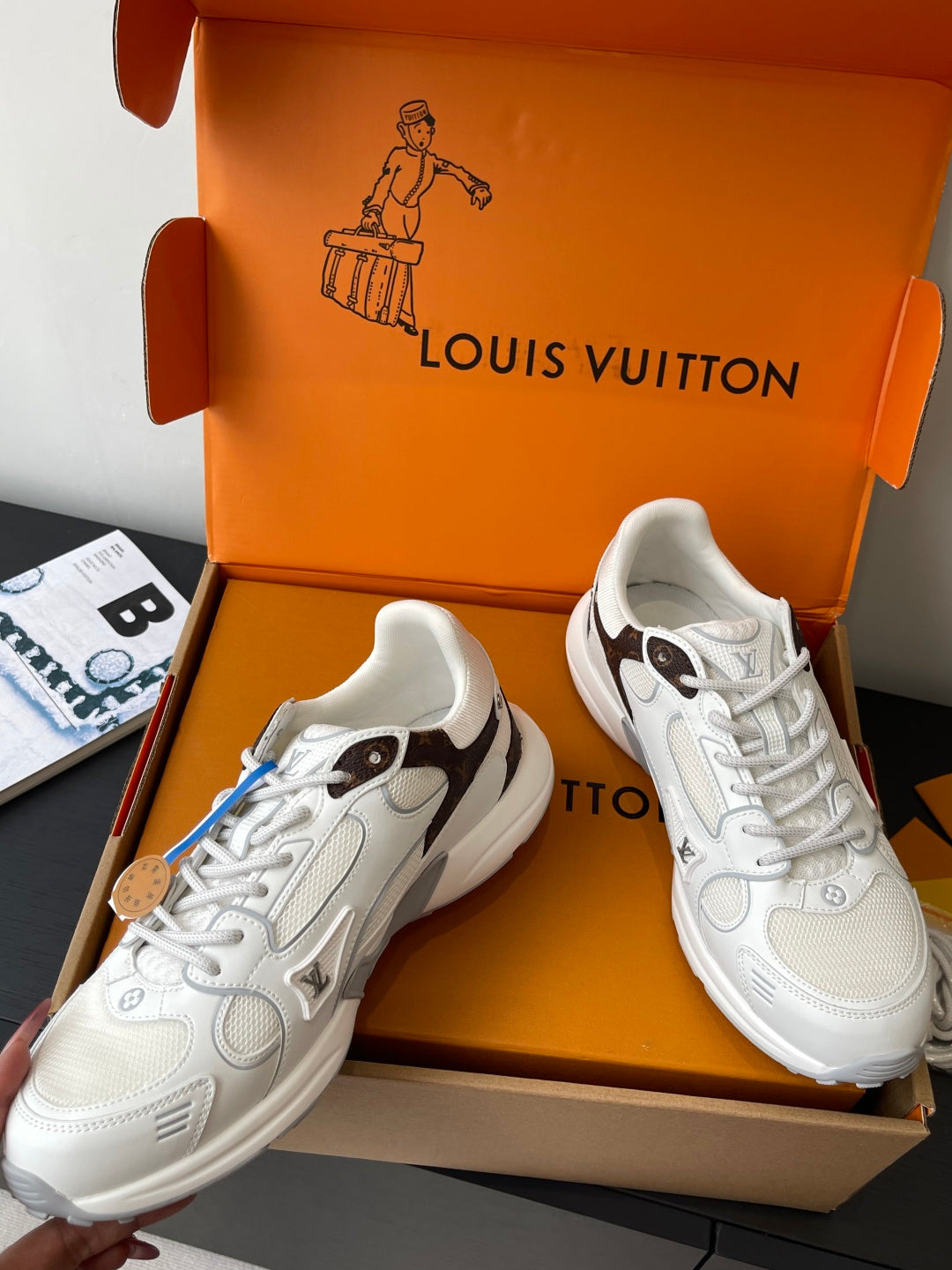 Pair of white lace-up sneakers in an open orange box, angled view with laces visible.