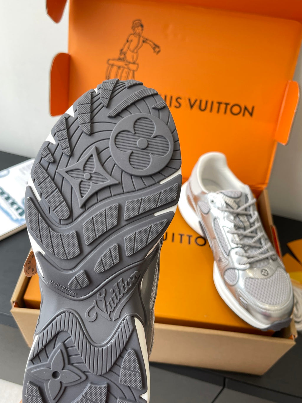 Shoe outsole view: gray textured tread on sneaker, with matching pair in box.