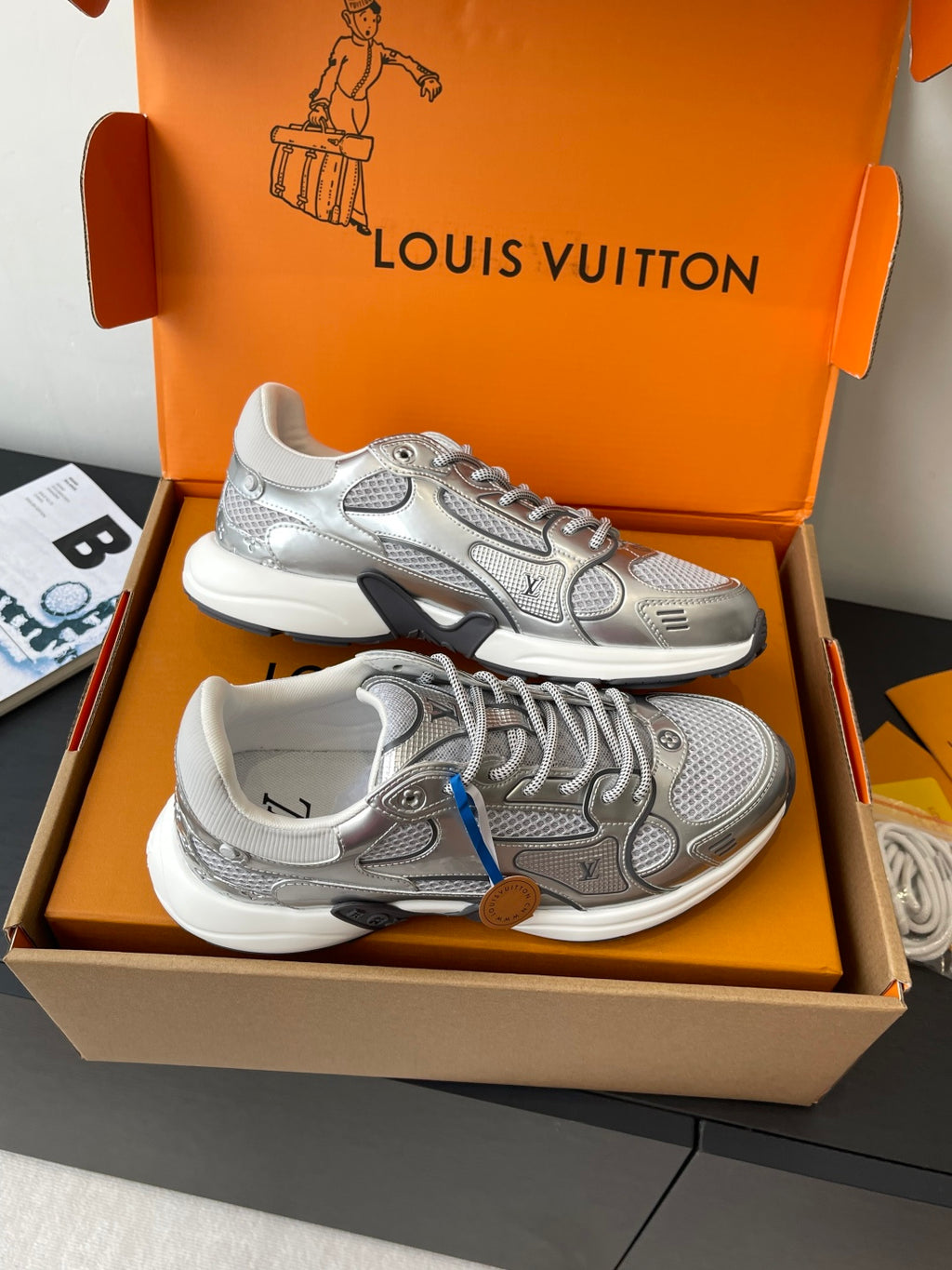 Front view of silver lace-up sneakers with logo panels, in orange gift box.