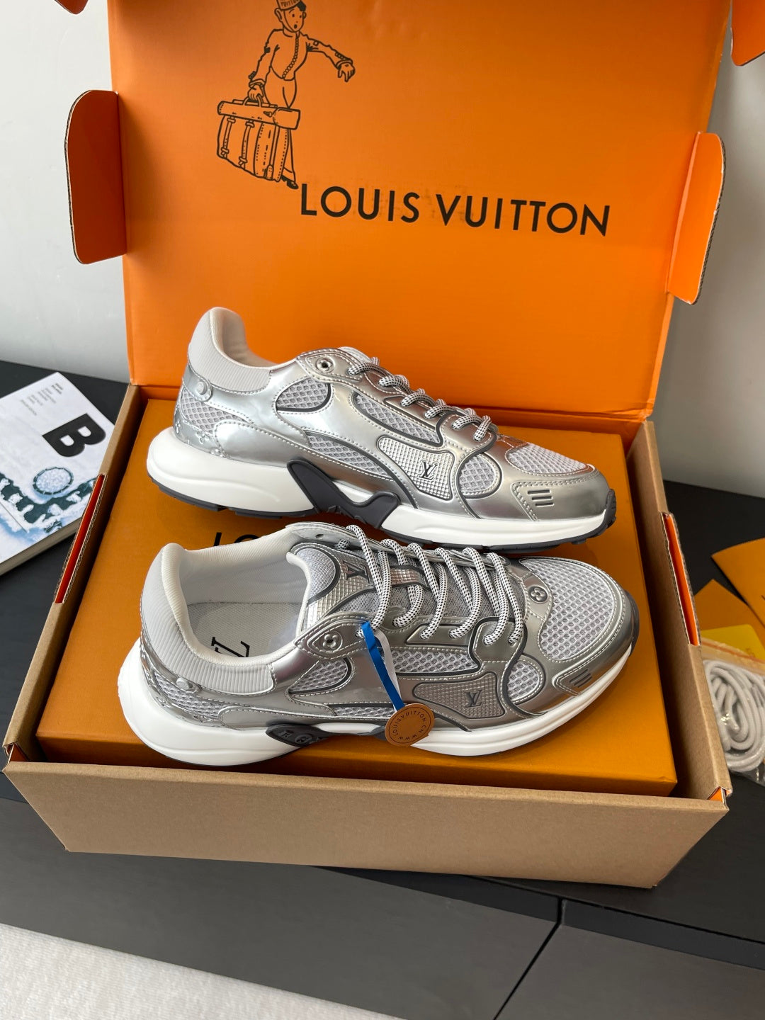 Front view of silver lace-up sneakers with logo panels, in orange gift box.