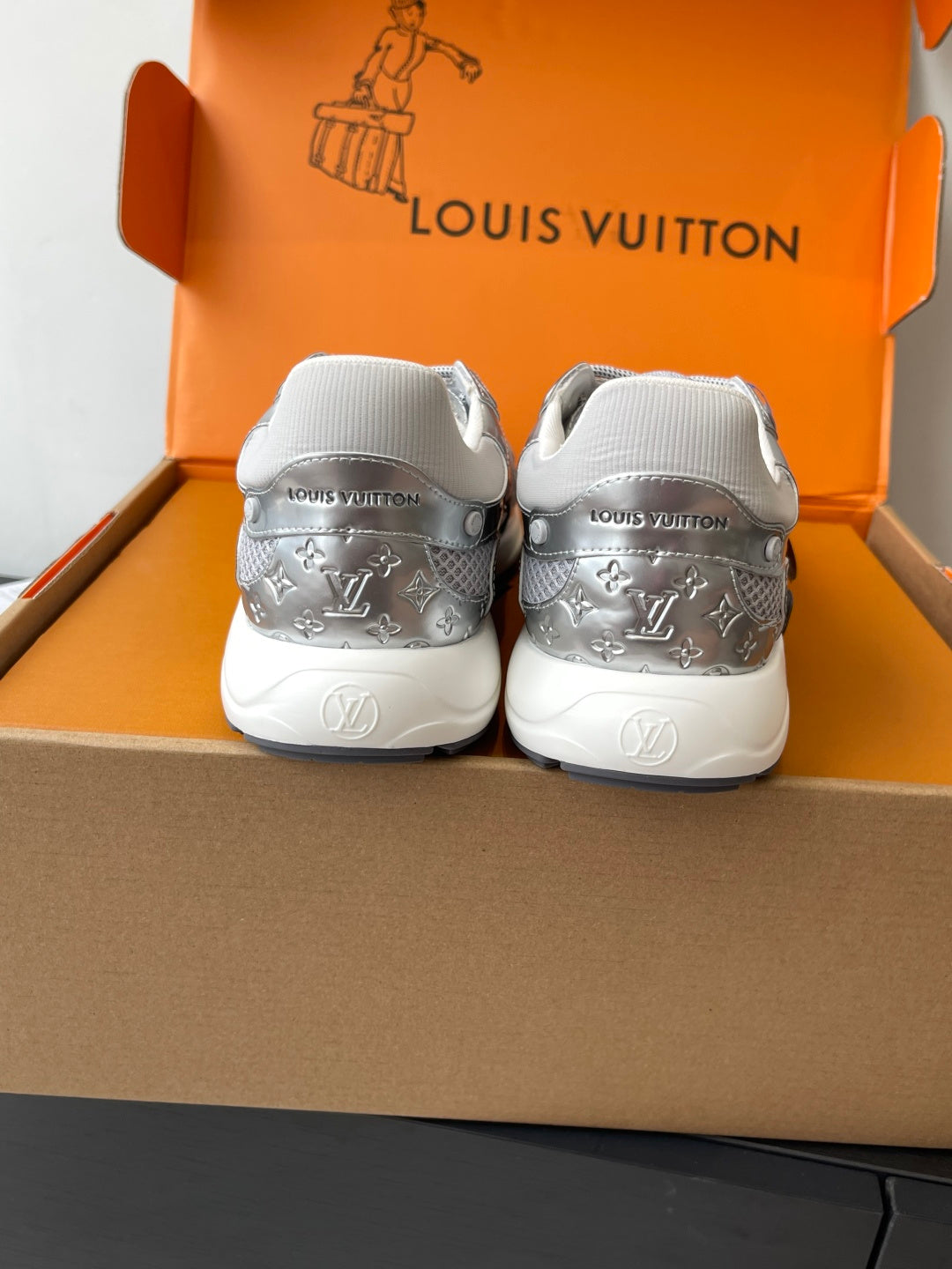 Back view of silver sneakers with heel branding, placed in orange box.