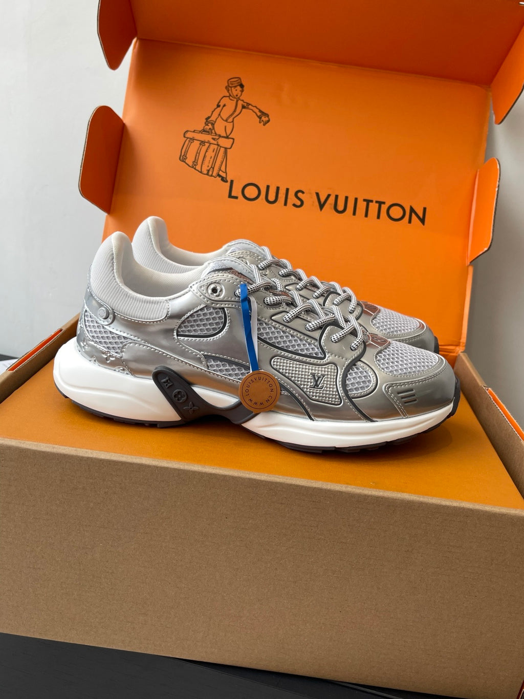 Silver lace-up sneaker with blue side stripe and logo details in orange box.