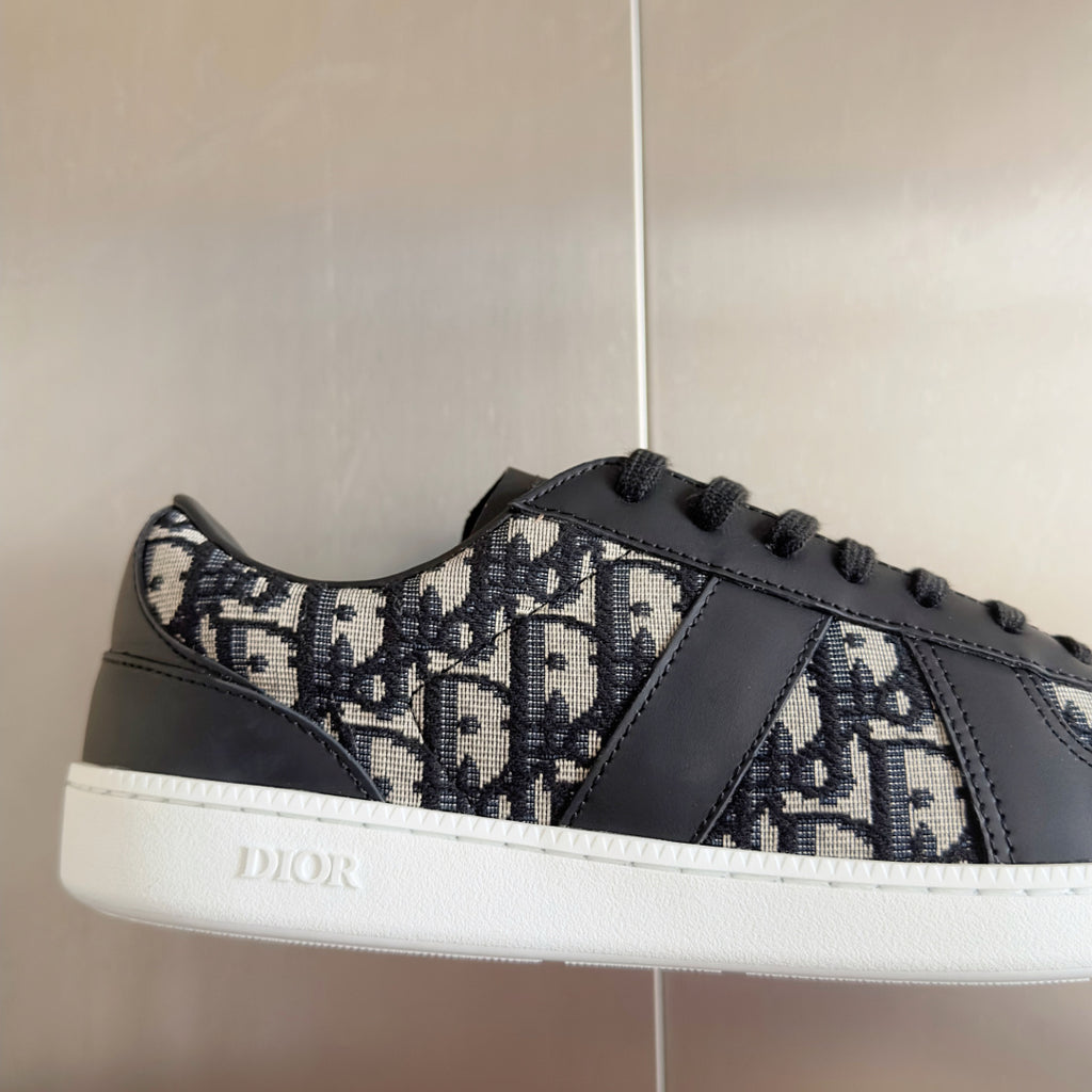 Low-top sneaker close-up, Dior monogram fabric with dark gray leather trim, black laces.