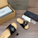 Black slide sandals shown from side, with buckle straps and cork-look sole