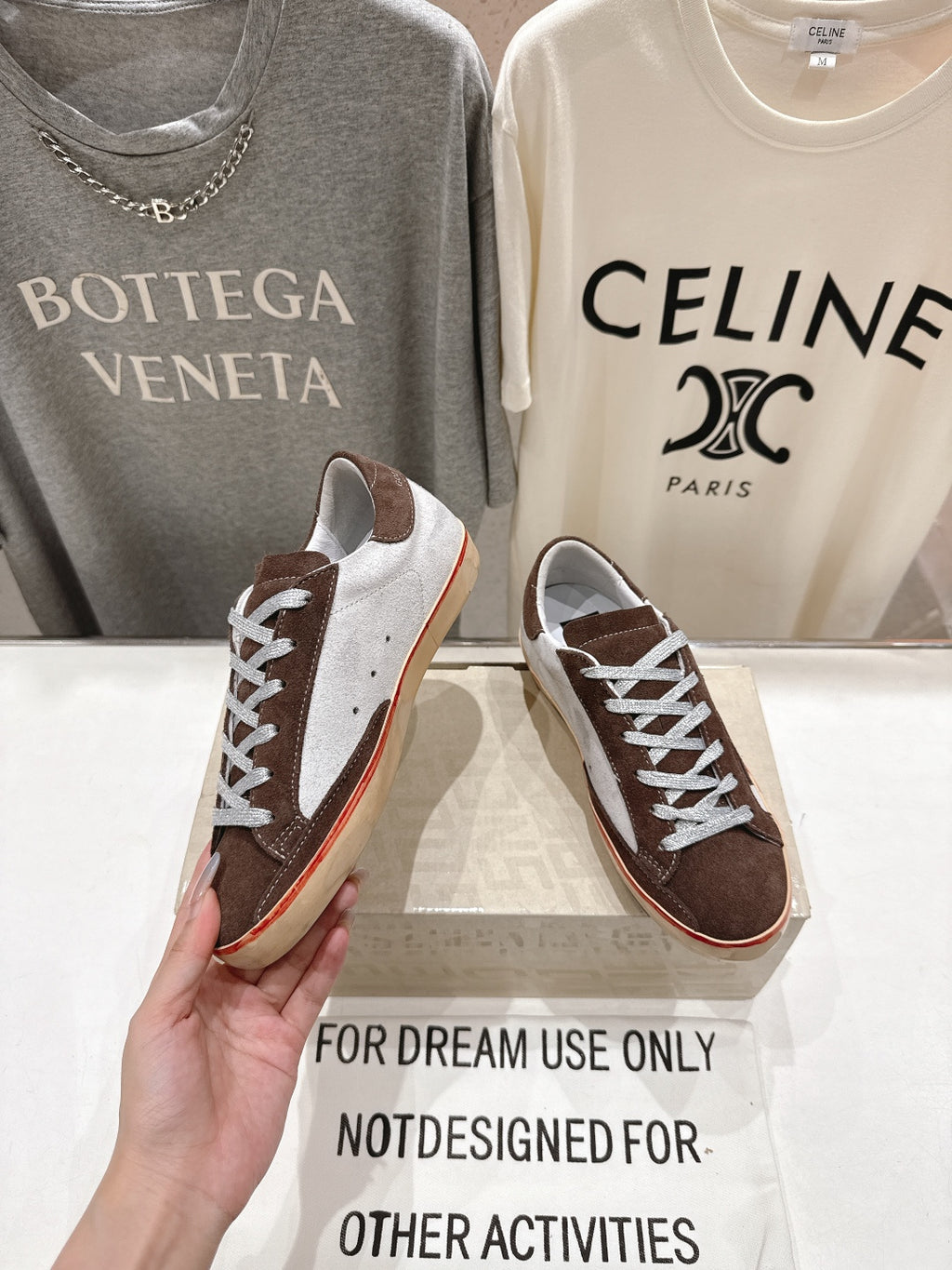 Hand holding brown and white lace-up sneakers with cream soles and red side stripe.