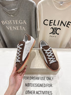 Hand holding brown and white lace-up sneakers with cream soles and red side stripe.