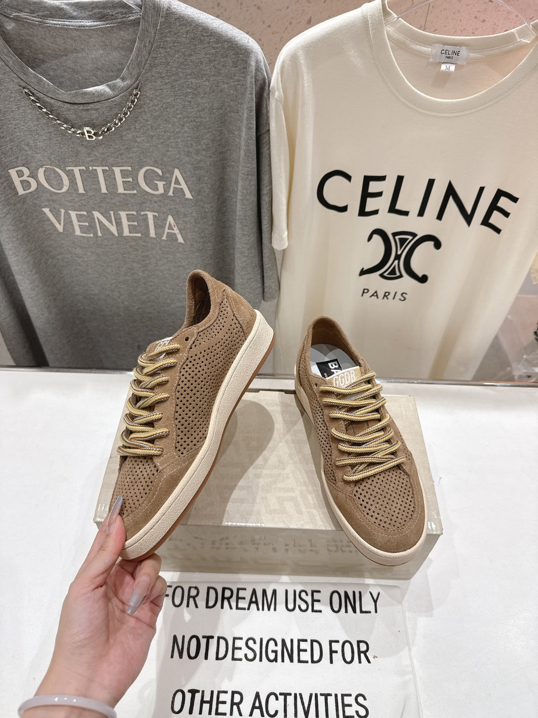 Tan low-top sneaker held near camera, perforated toe and gold lace-up