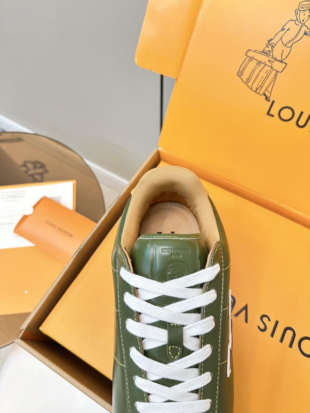 Green sneaker with white laces inside open orange box, heel tab visible