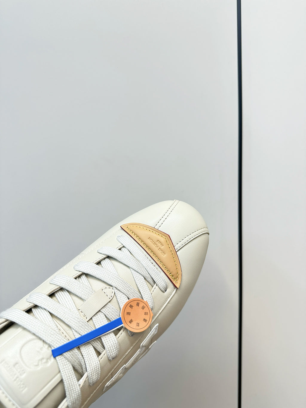 White leather sneaker toe close-up with tan panel, white laces, and blue lace detail