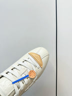White leather sneaker toe close-up with tan panel, white laces, and blue lace detail