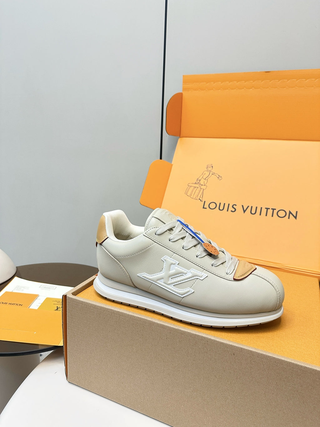 White leather sneaker on box with LV-style side logo, tan lace tab and blue lace strip