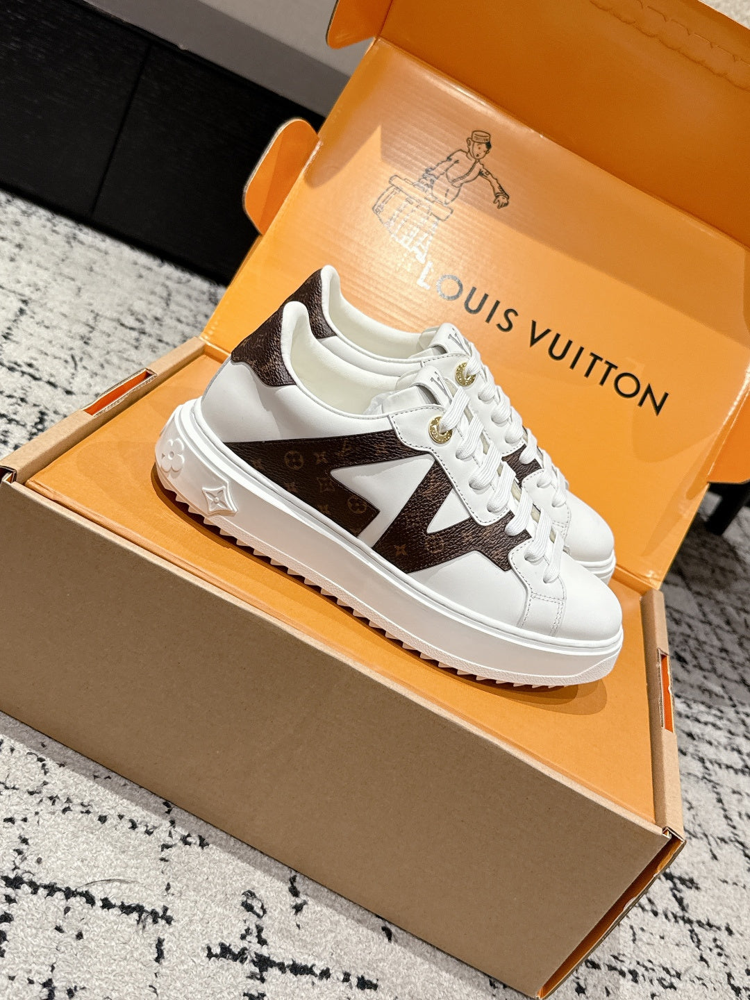 White low-top sneakers in an open box, brown star-like panel on the side