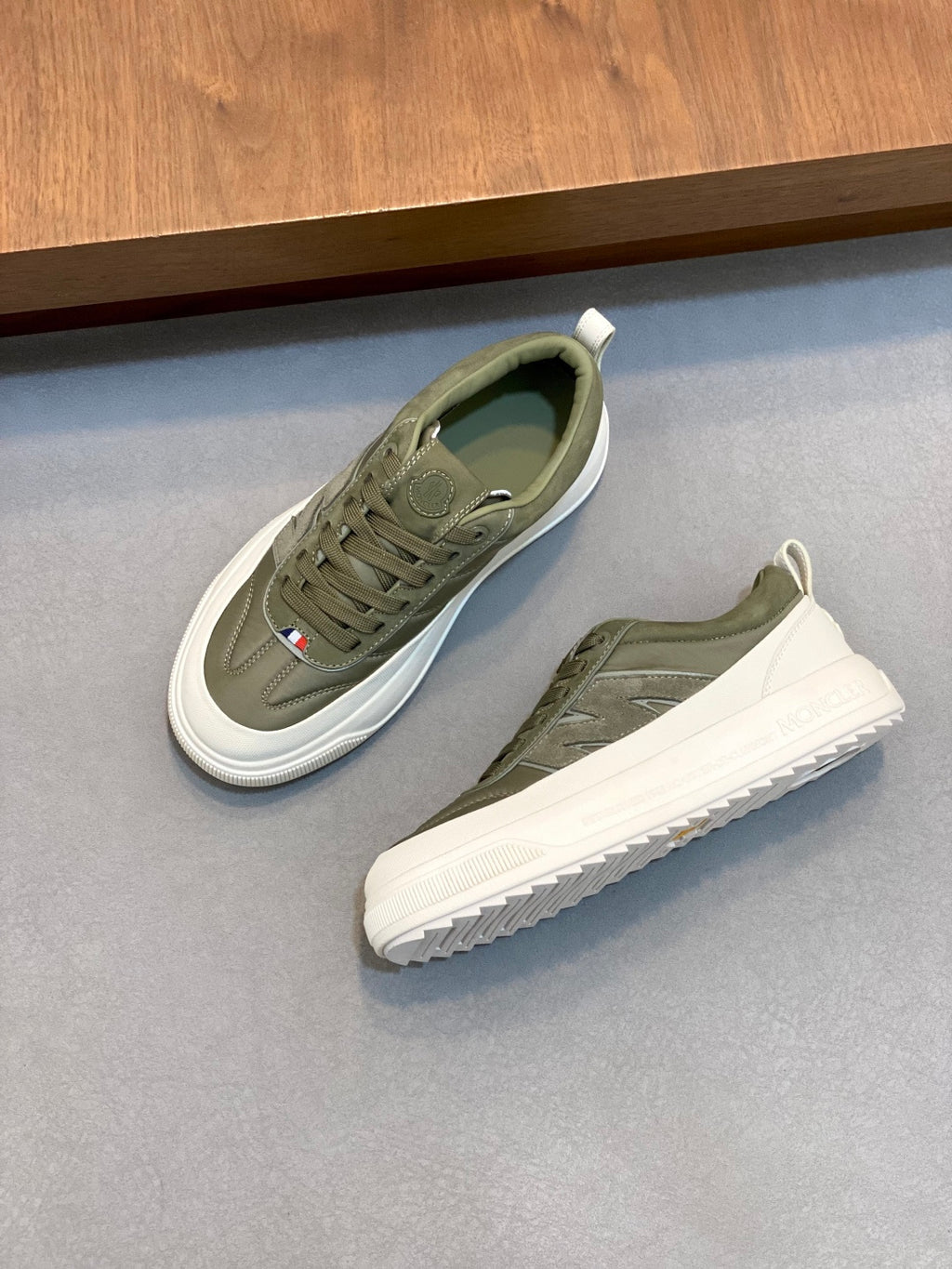 Pair of olive sneakers with white soles, lace-up style, and small side flag patch