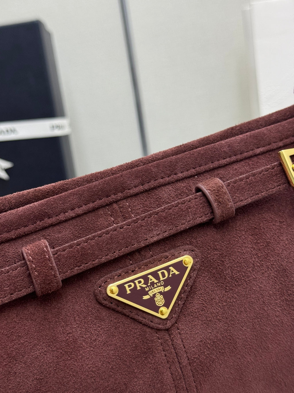 Dark red suede flap with gold-edged triangular logo plaque reading PRADA.