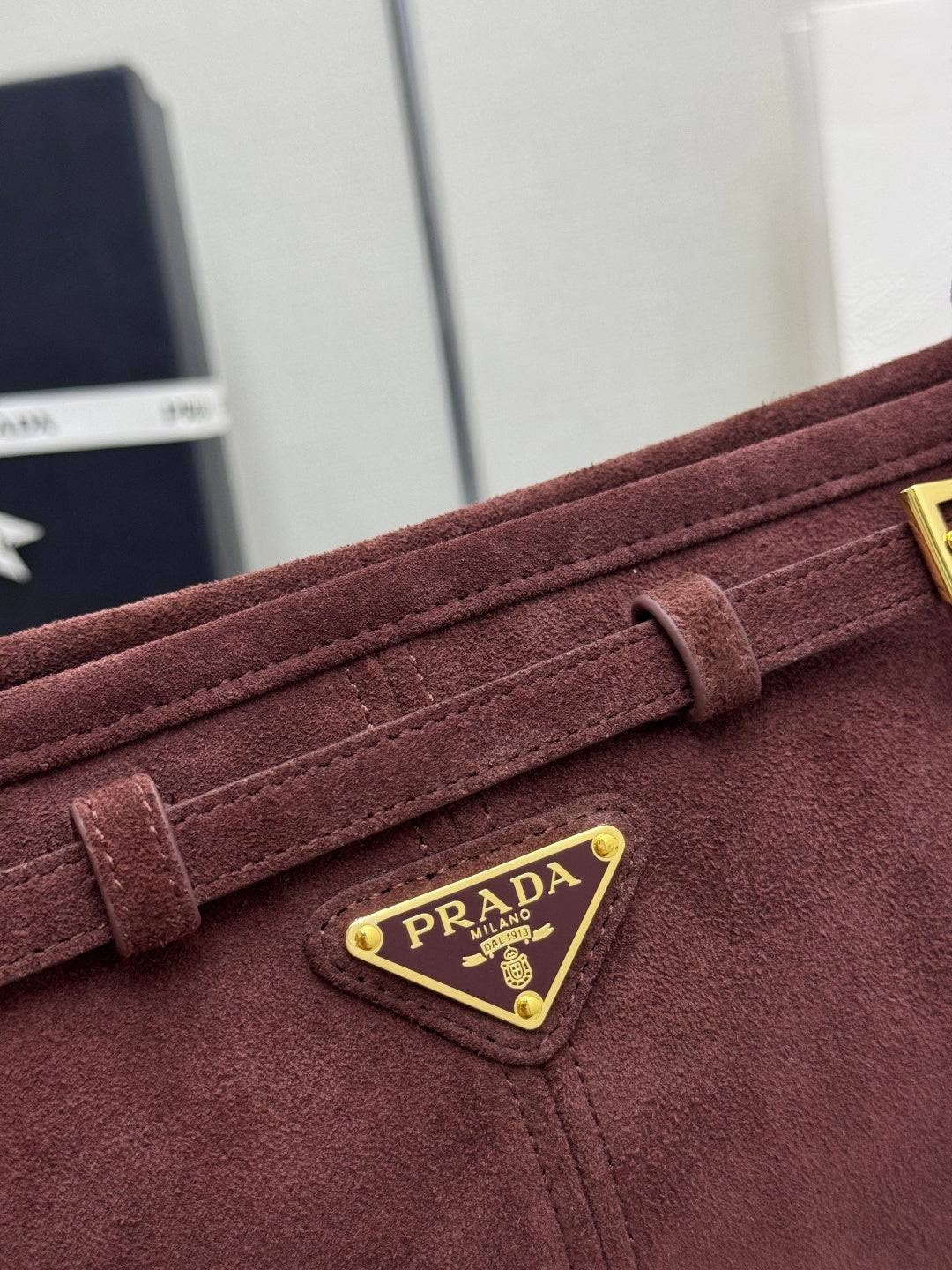 Dark red suede flap with gold-edged triangular logo plaque reading PRADA.