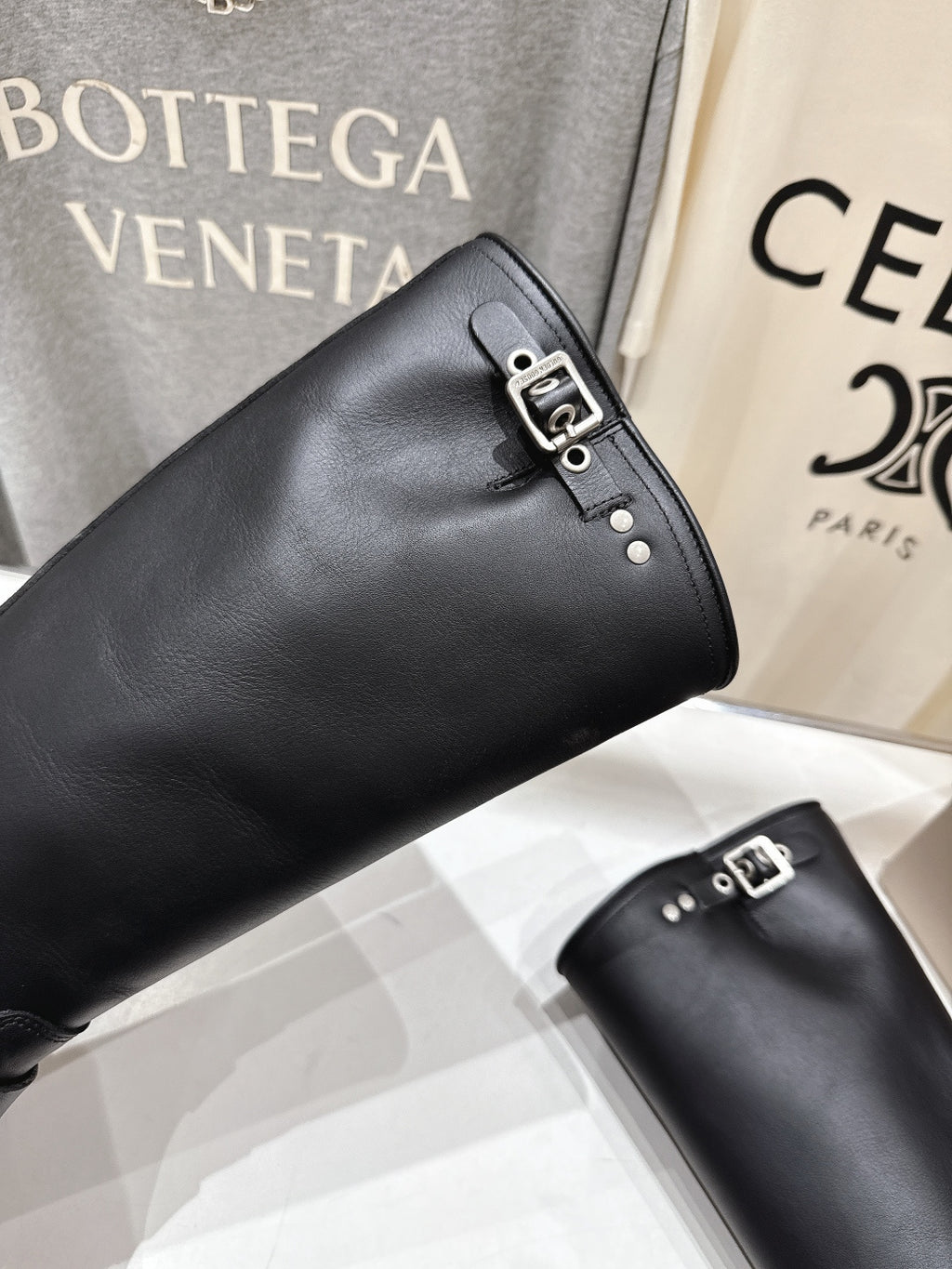 Black tall boots with metal buckle strap; angled close-up on shelf