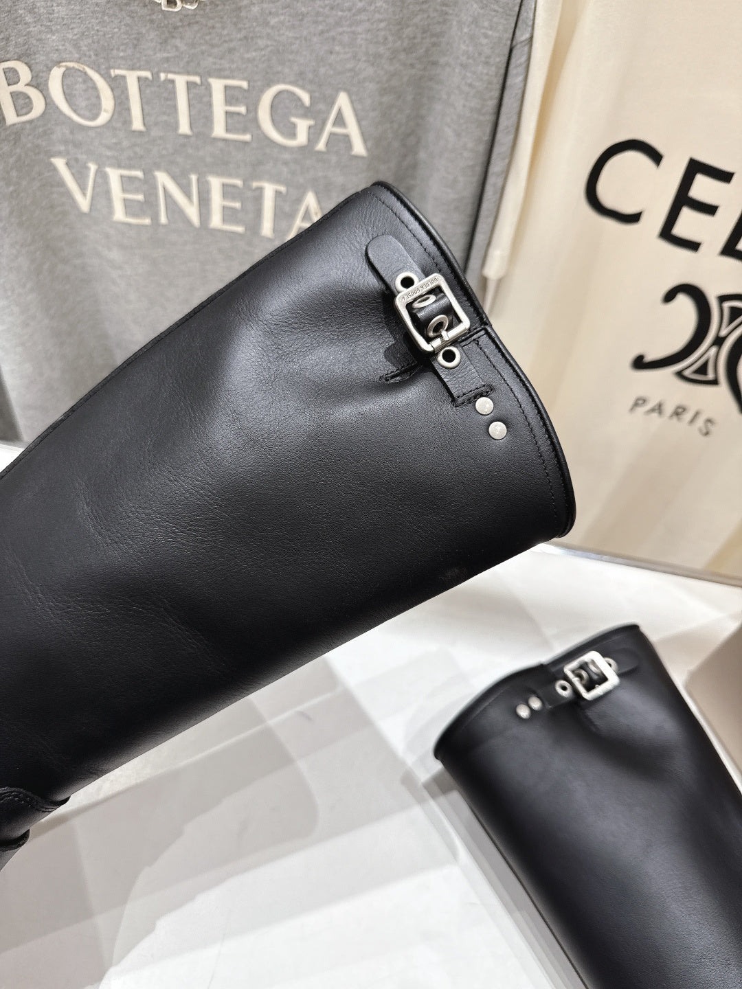 Black tall boots with metal buckle strap; angled close-up on shelf