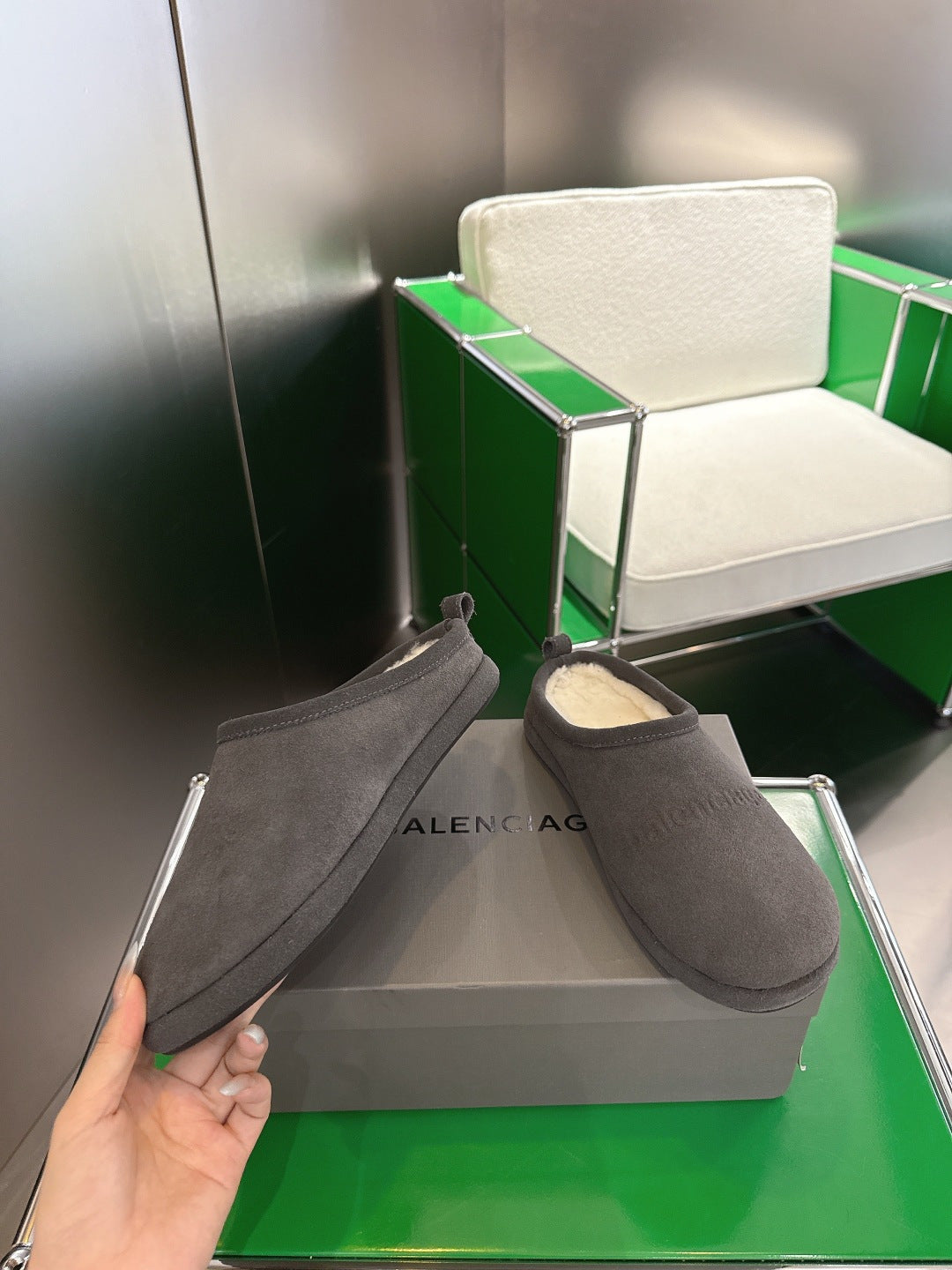 Dark gray suede slide sandals with plush cream lining, one shoe held near logo