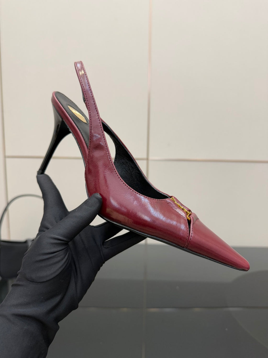 Burgundy red pointed-toe stiletto pump with gold emblem, close-up held by gloved hand