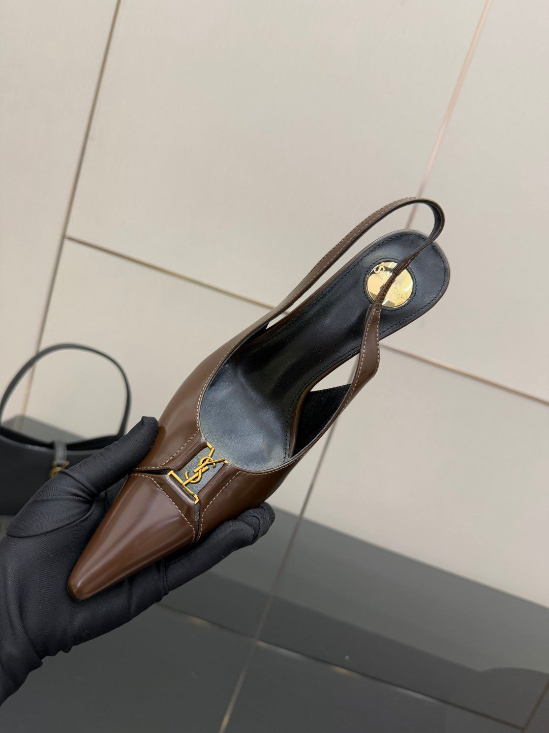 Brown pointed-toe slingback pump held by black glove, gold logo on strap