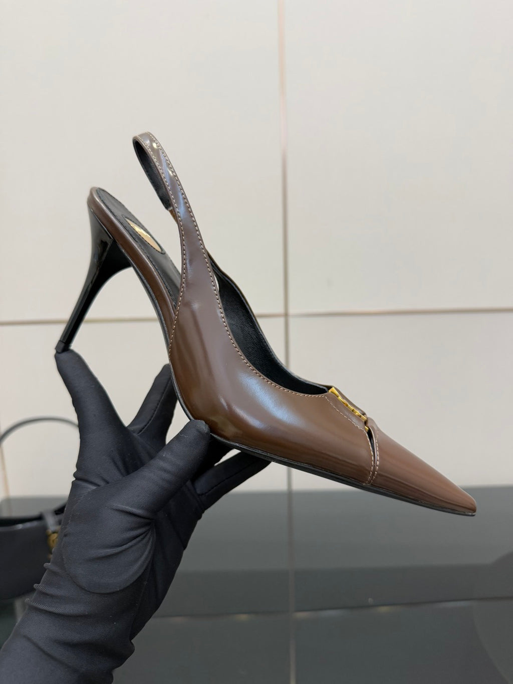 Brown leather pointed-toe slingback pump held in black glove, gold trim visible