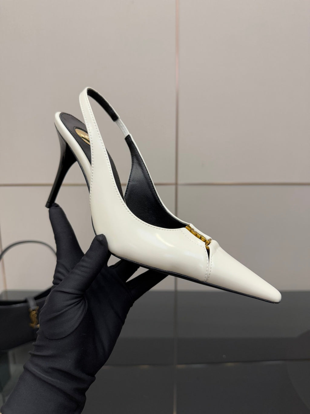 White pointed-toe slingback stiletto heel shoe held in a black glove, gold logo detail.