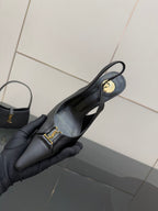 Black slingback stiletto pump with gold logo medallion, held in a gloved hand.