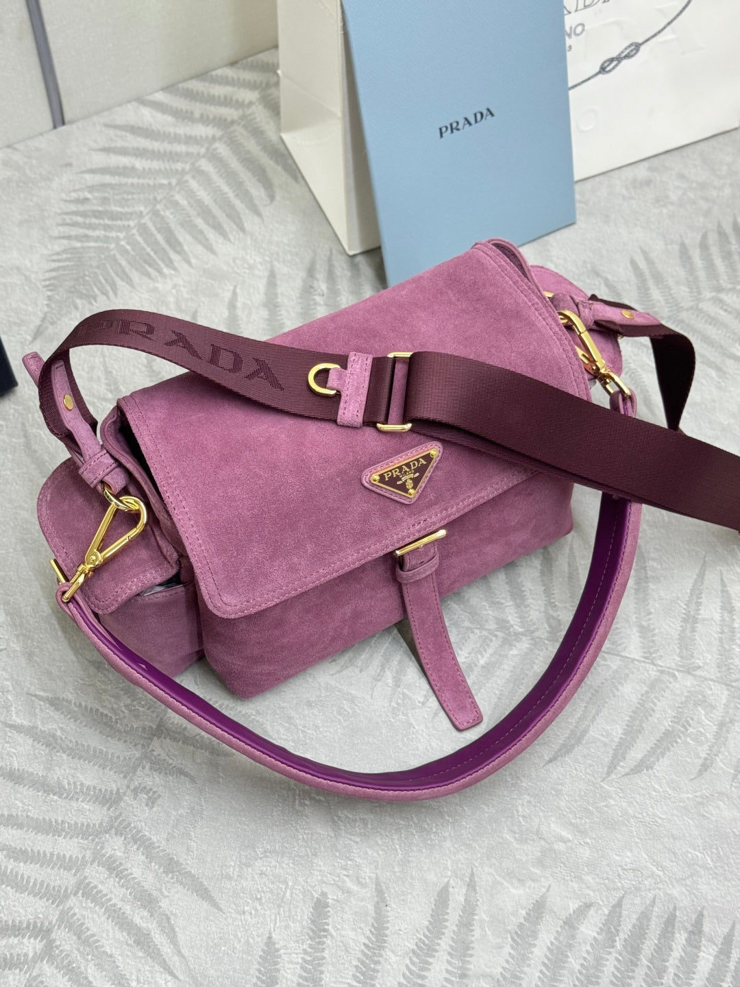 Purple suede shoulder bag lying sideways with front flap, strap, and gold-tone details