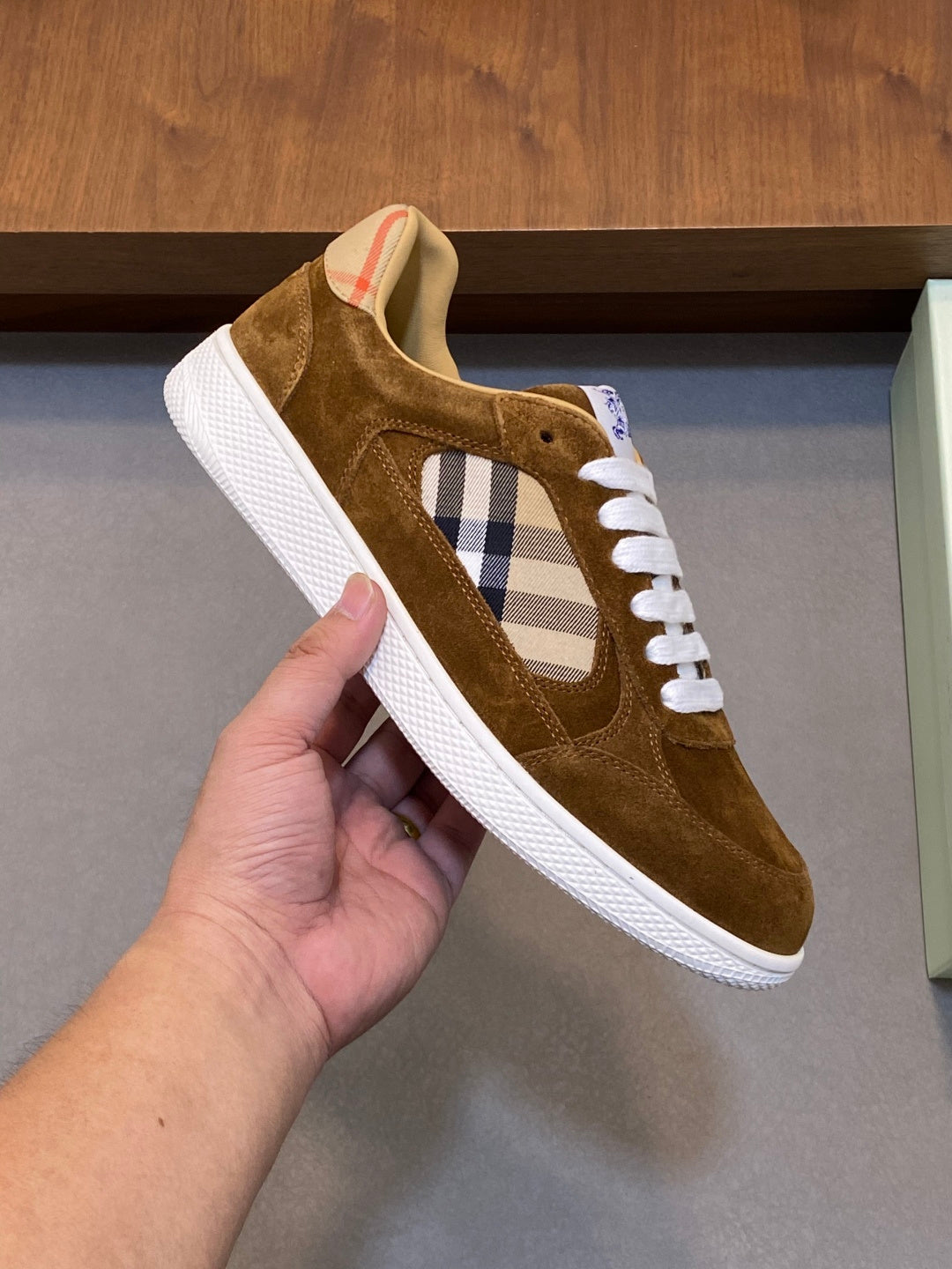 Brown suede sneaker held in hand, showing check panel, white laces, and white rubber sole