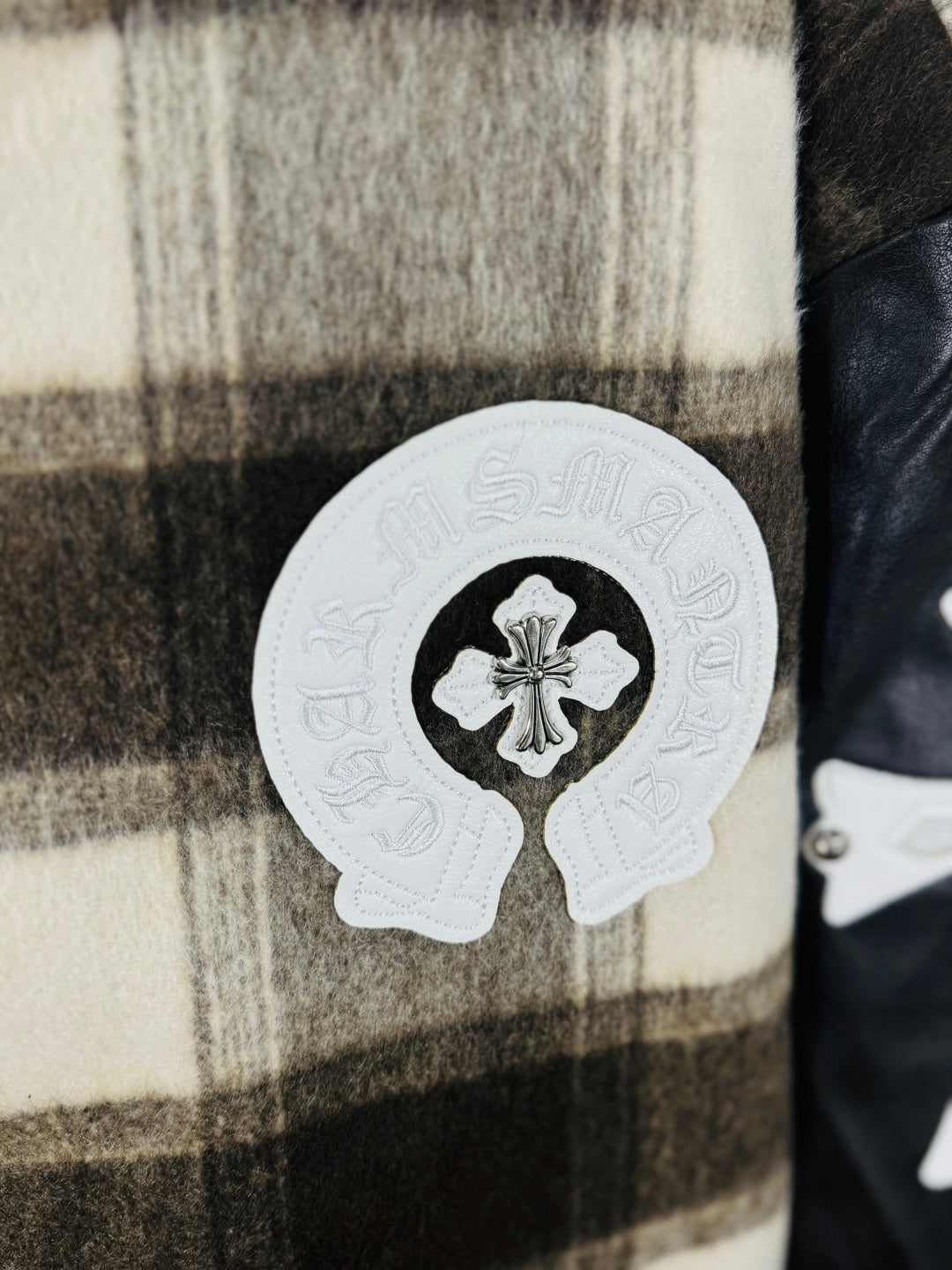 Close-up of plaid varsity jacket chest patch with white cross emblem on black-and-cream checks