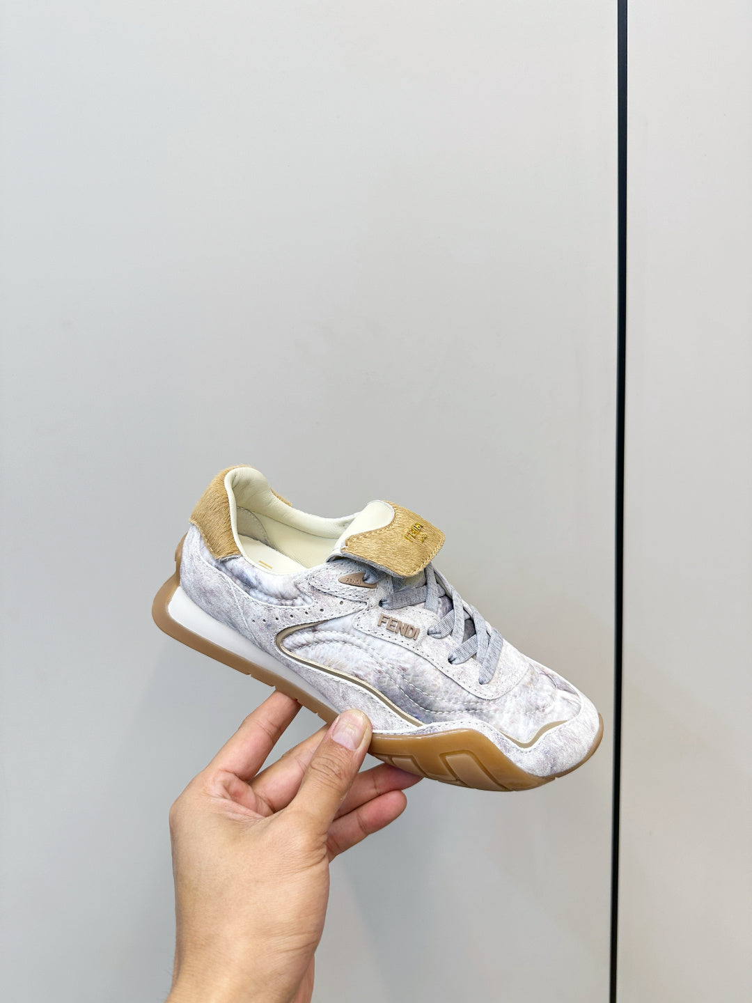 Fendi unisex sneakers held in hand, light gray with tan suede strap and lace-up.