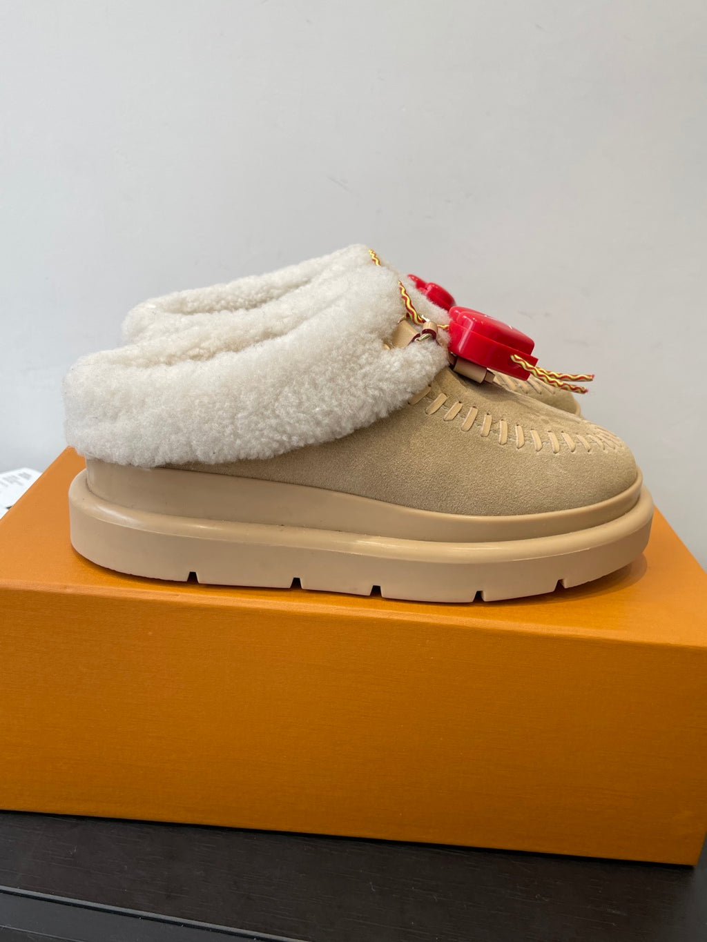 Tan slip-on shoe with thick cream shearling collar and red logo charm on strap