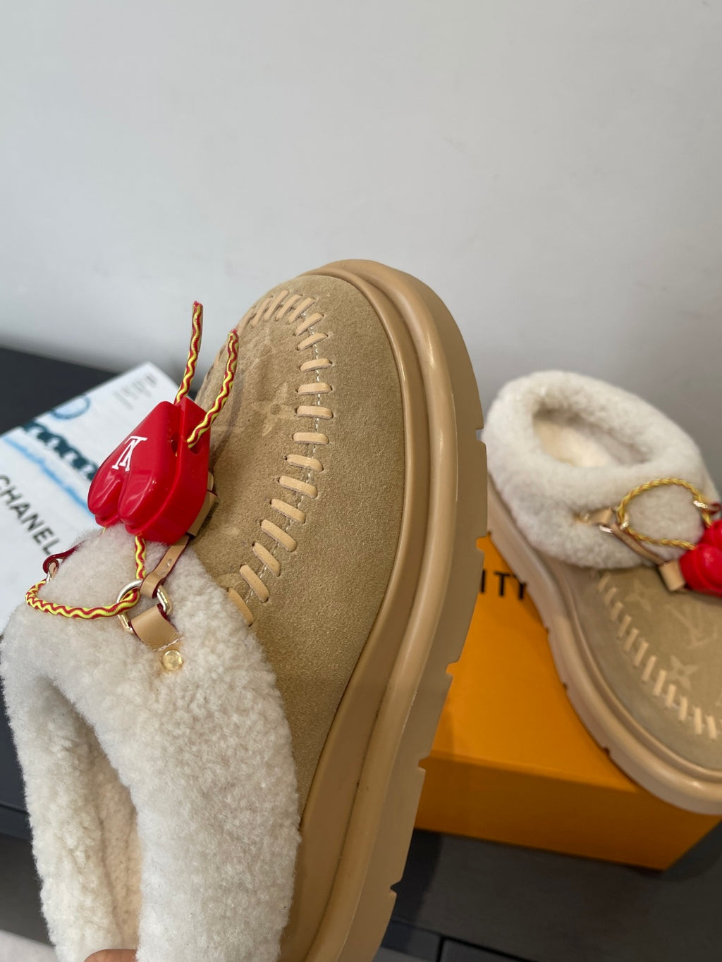 Tan suede slip-on shoes with shearling trim, red heart charm, and gold chain lace detail