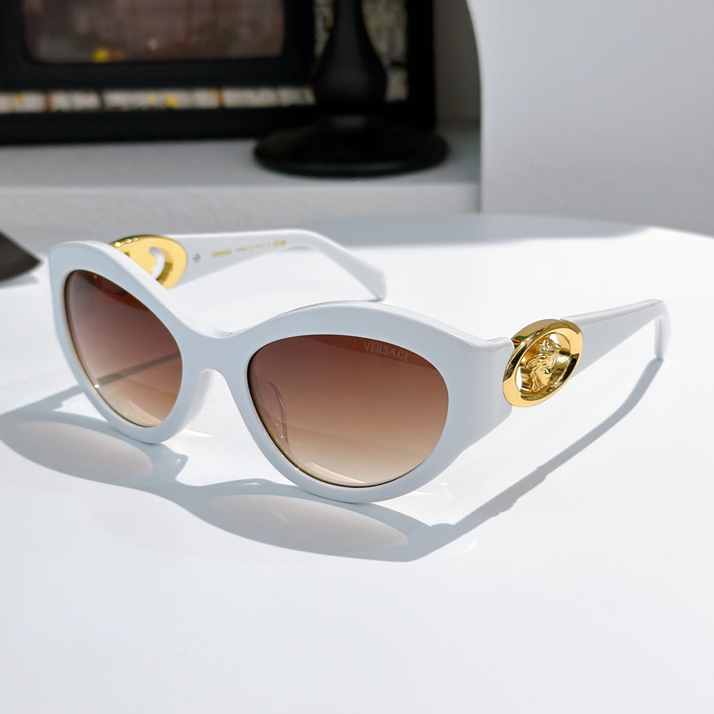 White cat-eye sunglasses with gold medallion logo detail and warm brown tinted lenses