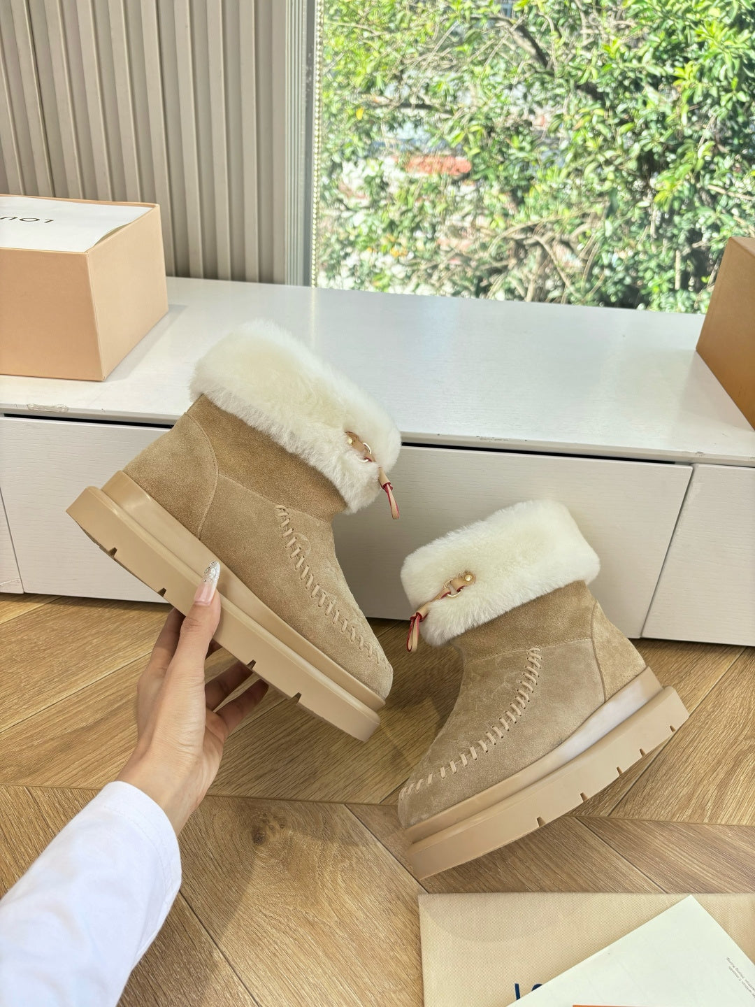 Tan suede ankle boots with white shearling cuffs and side emblem, held in hand