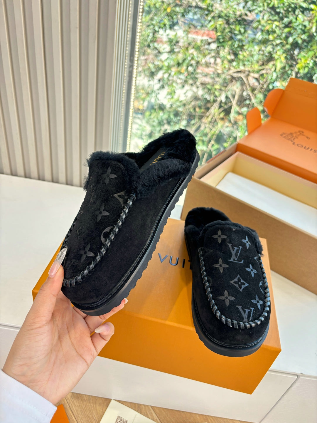 Black Louis Vuitton monogram slippers, one held and one resting on an orange box