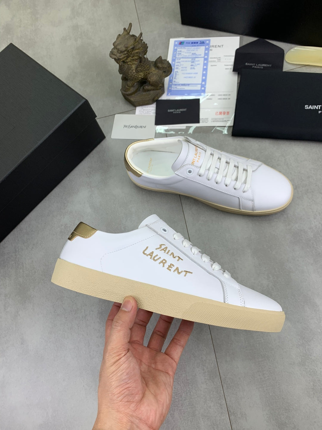 YSL unisex sneakers, white leather with tan gum sole and side logo, held in hand.