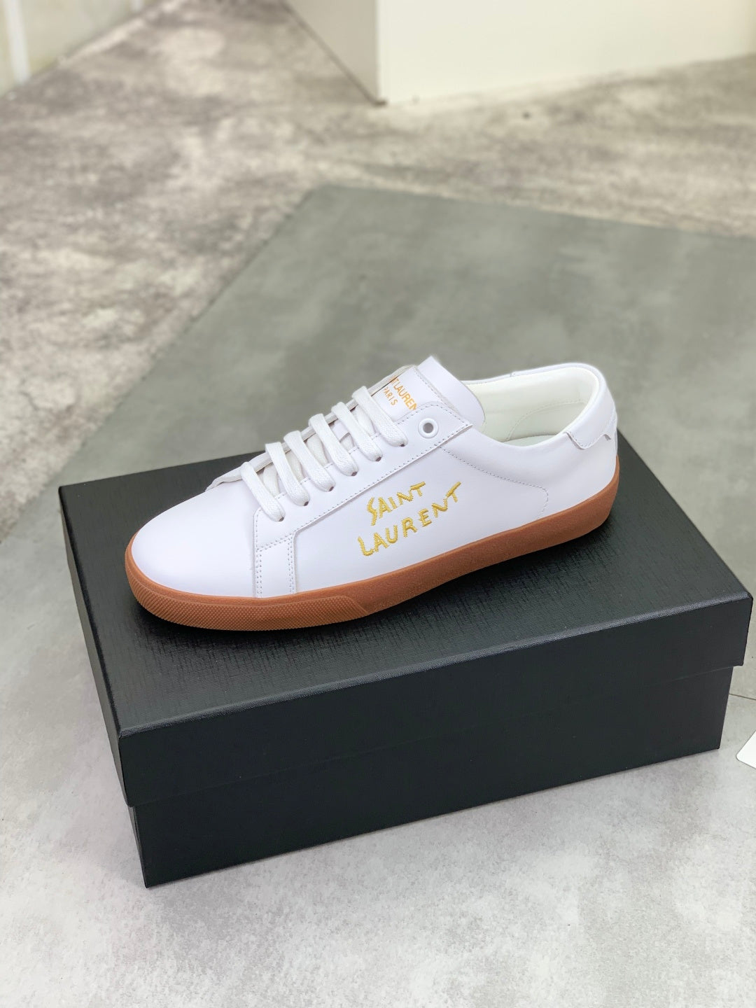 White low-top sneaker with gold logo on side and tan rubber sole