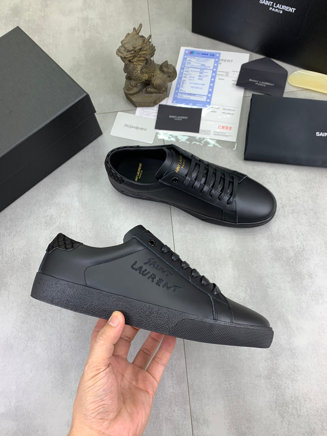 Dark leather lace-up sneakers held in hand, showing side logo and flat sole.