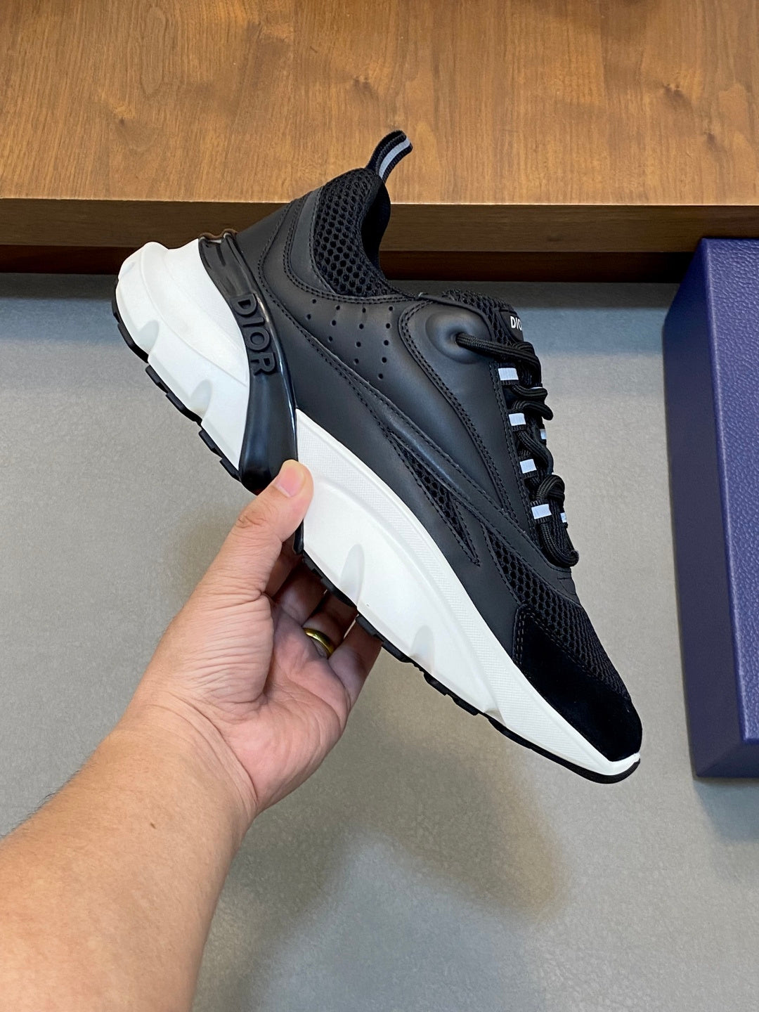 Black and dark gray Dior sneakers with white sole, held by hand, lace-up style