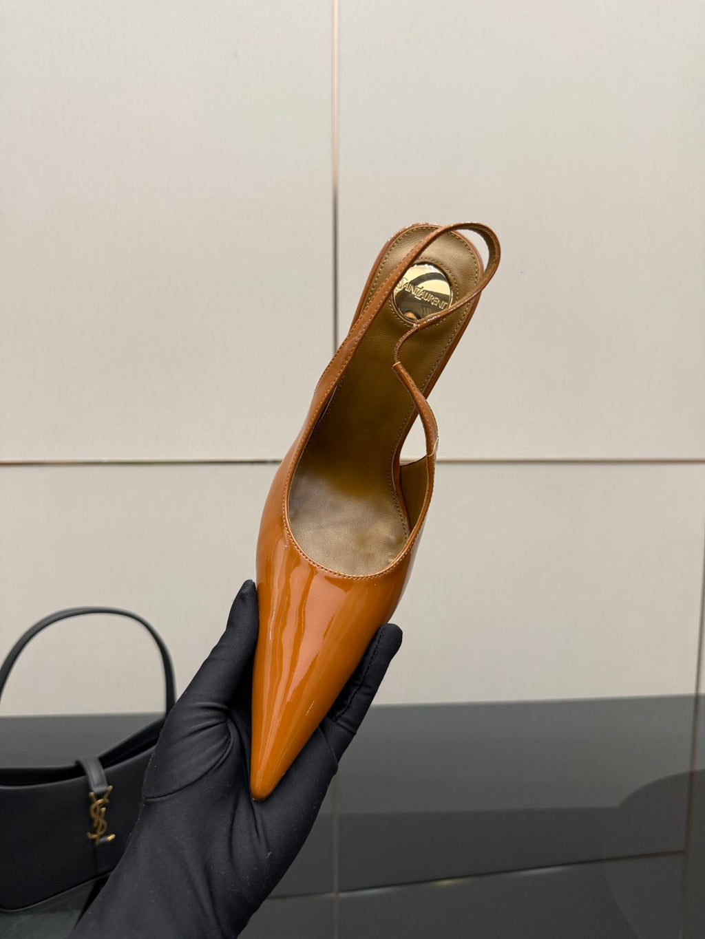 Tan-orange patent pointed-toe slingback high heel, held in black-gloved hand