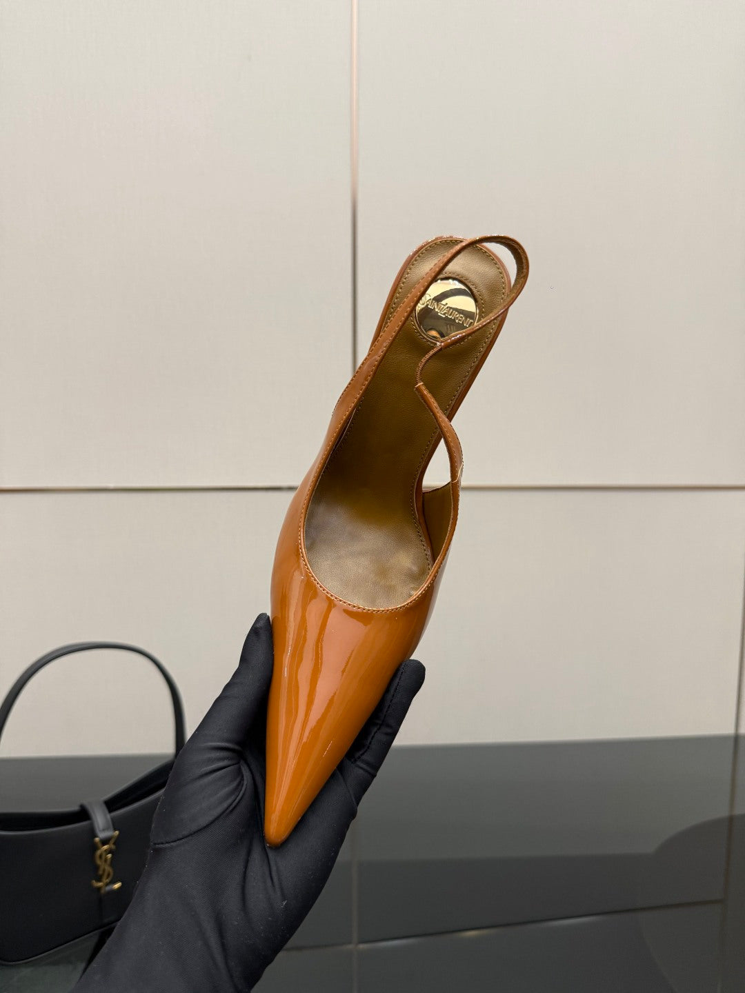 Tan-orange patent pointed-toe slingback high heel, held in black-gloved hand
