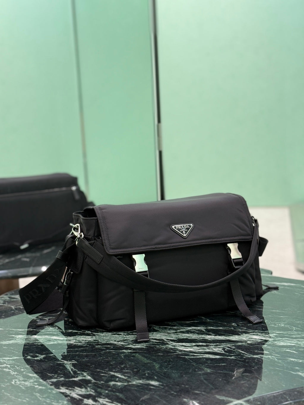 Black Prada messenger bag angled view, front flap with logo plate and buckled straps.