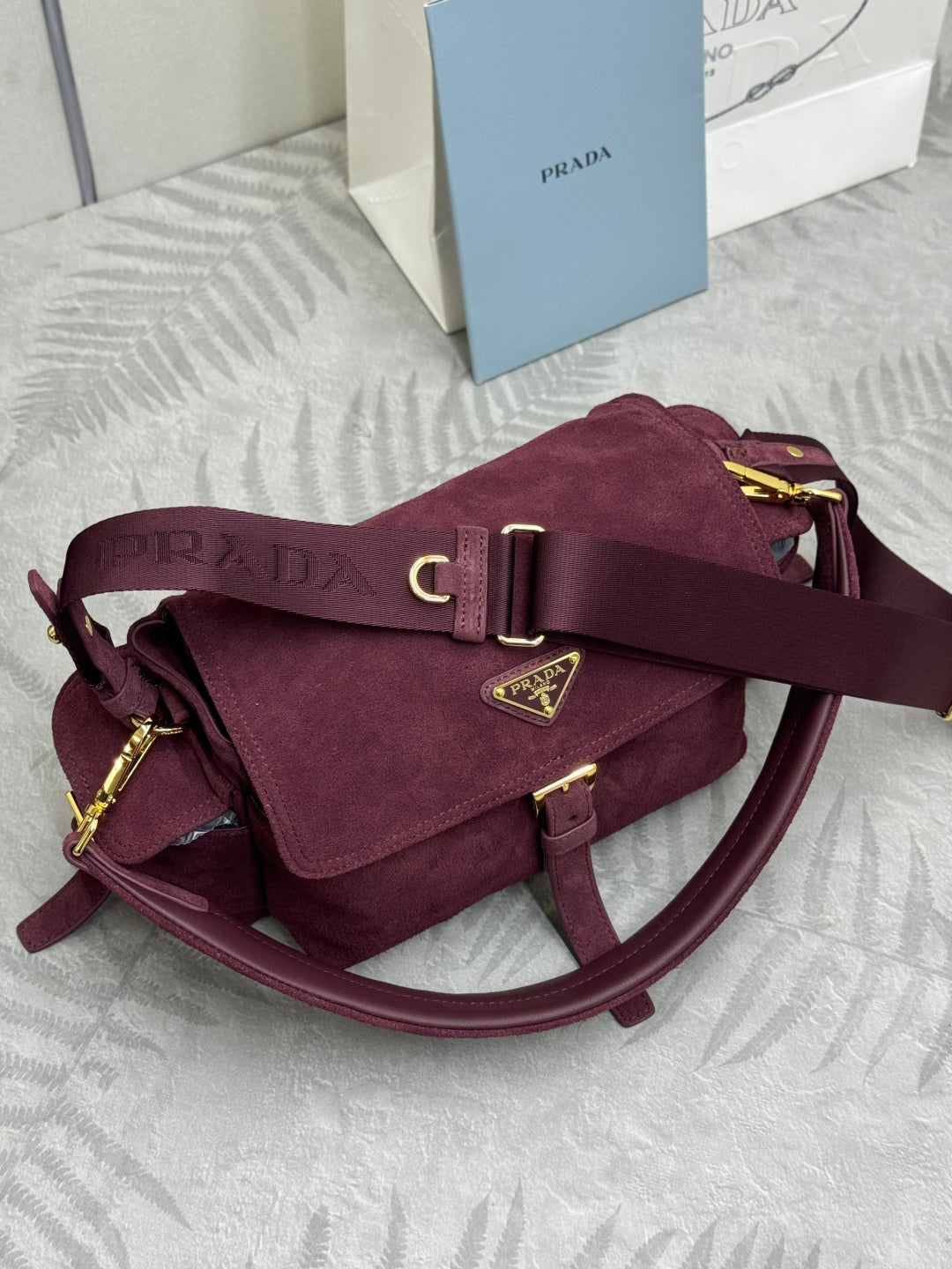 Burgundy suede Prada shoulder bag with front flap, gold buckle, and curved strap