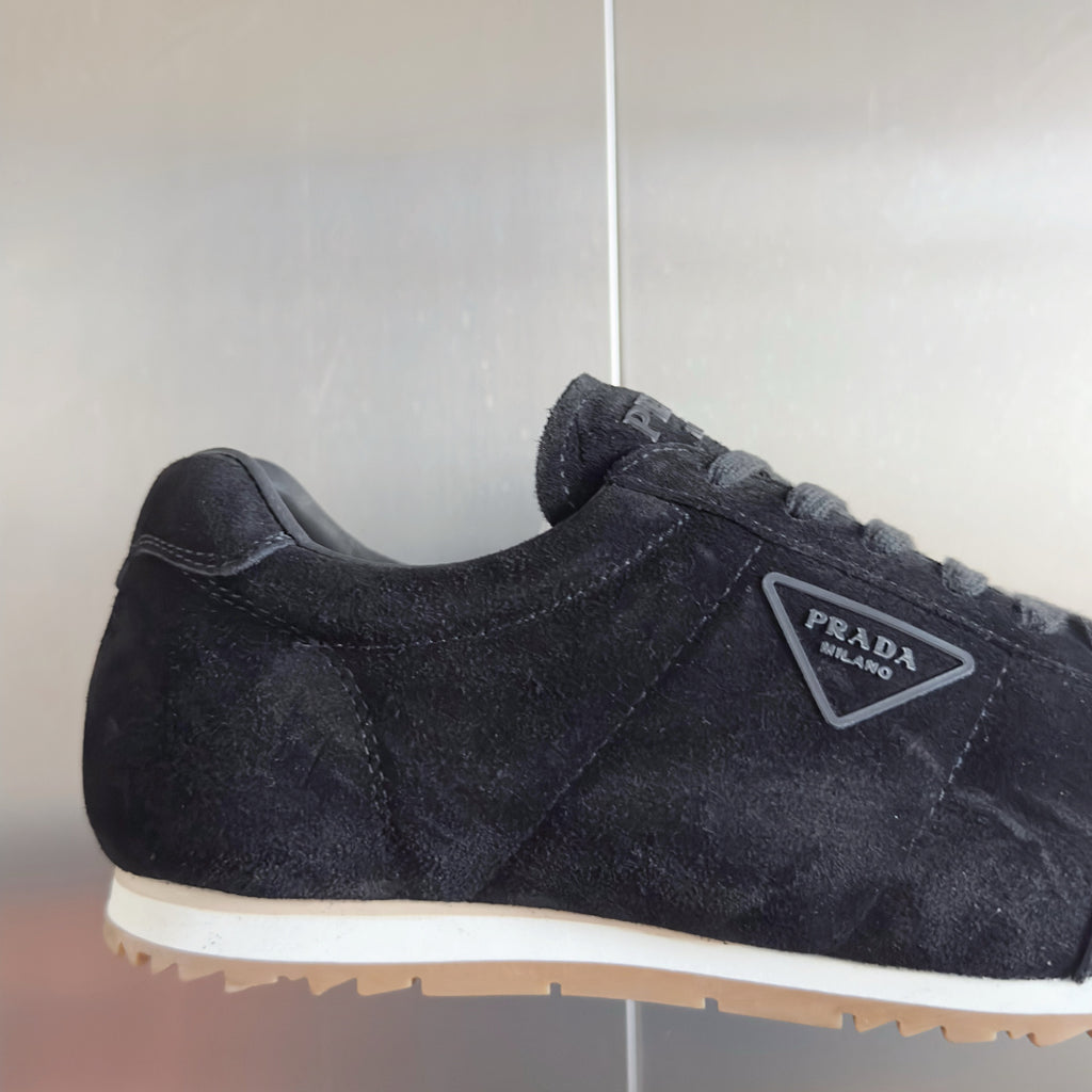 Black suede sneaker side view with Prada triangular logo patch and lace-up.