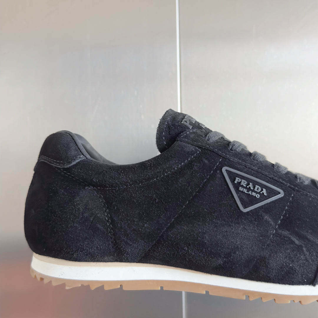 Black suede sneaker side view with Prada triangular logo patch and lace-up.