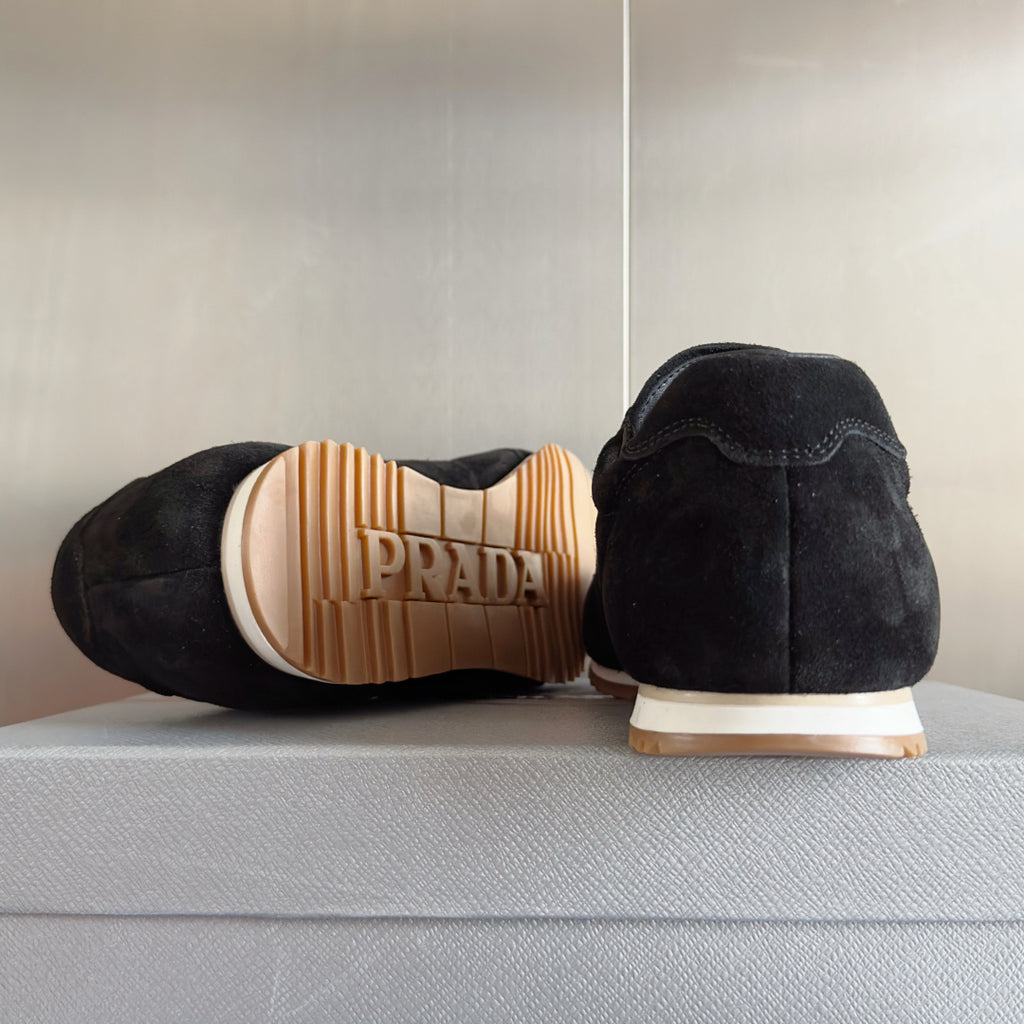 Black suede sneakers with rear stitching and PRADA on striped outsole tread.