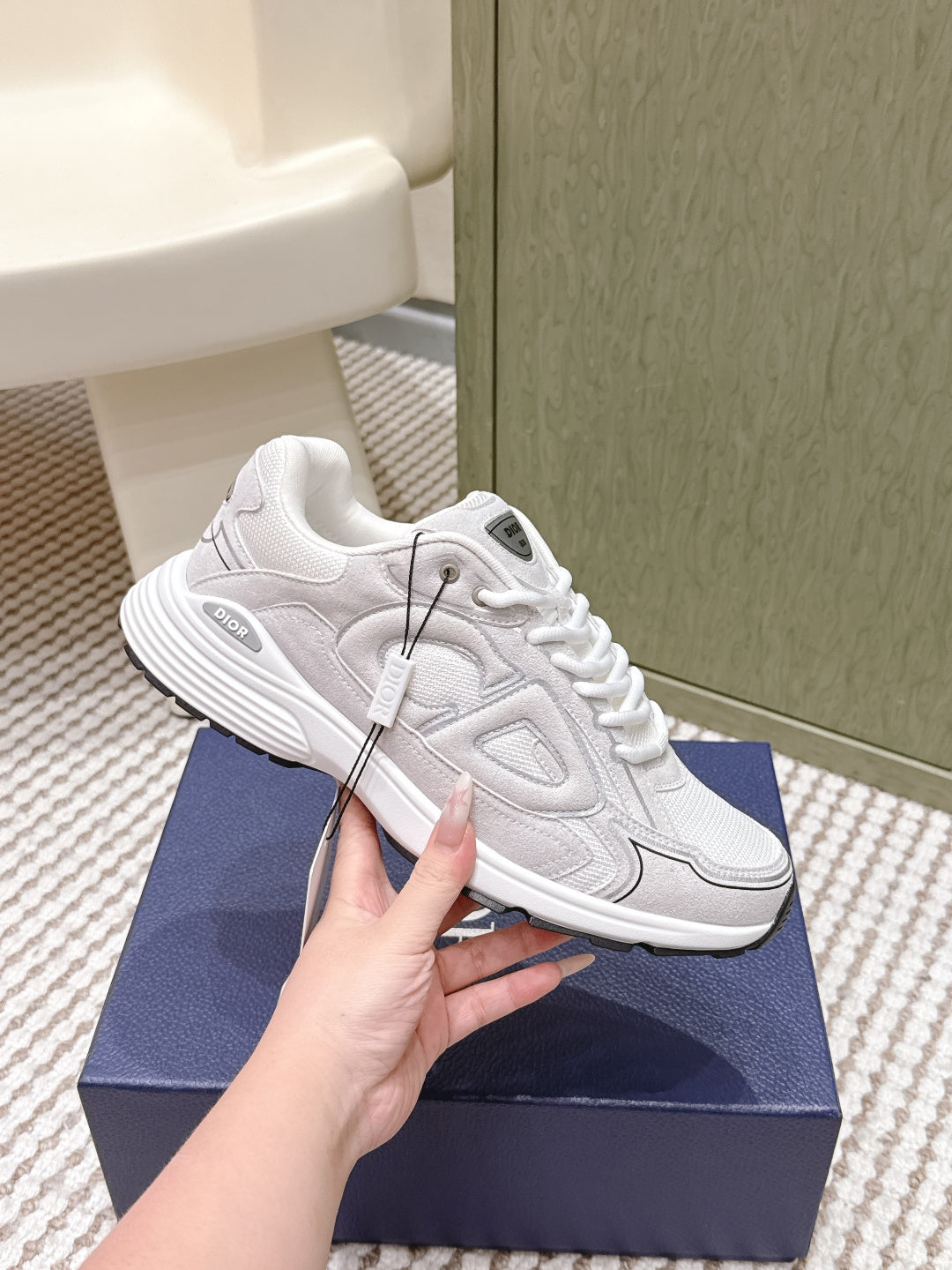 White Dior sneakers, angled side view, held by hand; textured panels and laces.