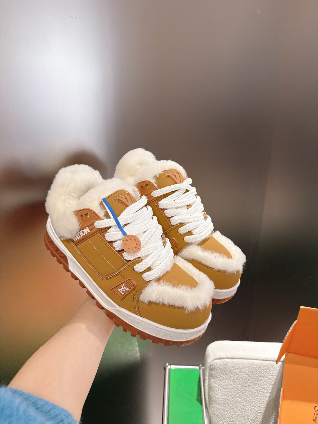 Unisex tan-and-white fur-trimmed sneakers with white laces, held above a shoebox